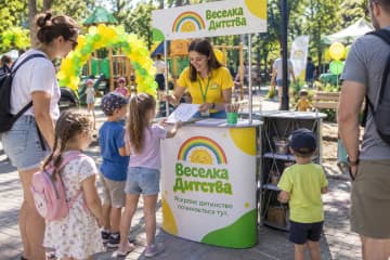 Промостіл Oval розміром 100×50 см у дитячій зоні сімейного фестивалю, де видно овальну стільницю та друковану панель спереду.