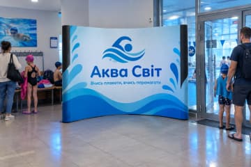 Криволінійна поп-ап стіна Pop-up Arc 400×230 см у лобі школи плавання біля входу до басейну з вертикальними друкованими ПВХ-панелями.