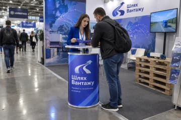 Телескопічний промо-стіл з LED-підсвіткою та друкованою передньою панеллю стоїть на логістичній виставці серед людей, палет і стендів.