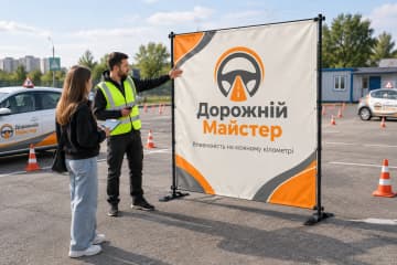 Брендвол Cross встановлено на майданчику автошколи Дорожній Майстер поруч із навчальним авто та конусами, і видно стійки на Х-подібних опорах.