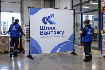 Брендвол Cross стоїть у лобі логістичної компанії Шлях Вантажу поряд із коробками та візком, і видно стійки на Х-подібних хрестовинах.